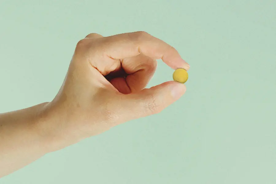 A white person’s hand pinching a yellow pill against a green background